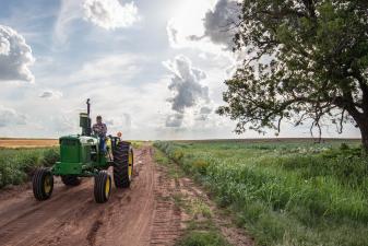 a green tractor drives along a dirt road with blue skies and lush grass